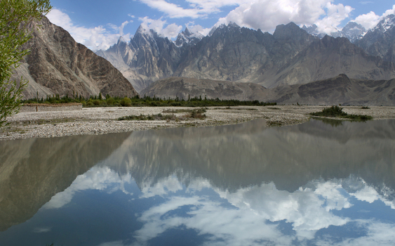 View of the K2 trek landscape in Hushe Pakistan featuring a calm river reflecting the dramatic snow capped peaks and clouds under a bright sky. View of the K2 trek landscape in Hushe Pakistan featuring a calm river reflecting the dramatic snow capped peaks and clouds under a bright sky.