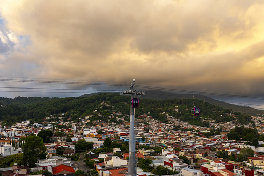 Aerial view of the Teleferico Uruapan cable car system gliding over the dense urban landscape with colorful rooftops and green hills under a dramatic sky in Uruapan Michoacan Mexico.