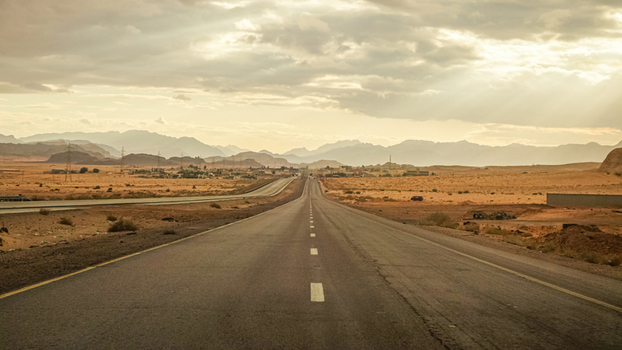View of a long asphalt road stretching through the arid desert landscape under a cloudy sky with soft golden light in Jerash Jerash Governorate Jordan. View of a long asphalt road stretching through the arid desert landscape under a cloudy sky with soft golden light in Jerash Jerash Governorate Jordan.