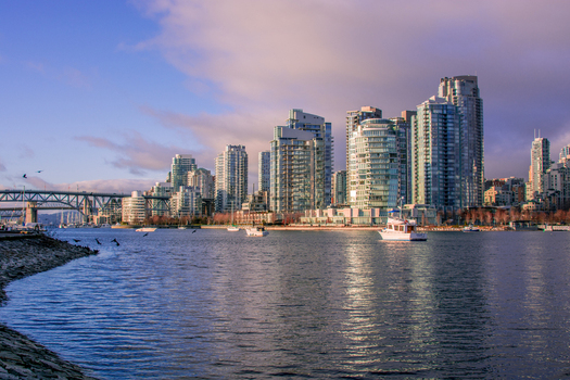 View of the Vancouver skyline across False Creek featuring modern glass skyscrapers the Burrard Street Bridge and boats on the water under a soft sunset sky Canada.