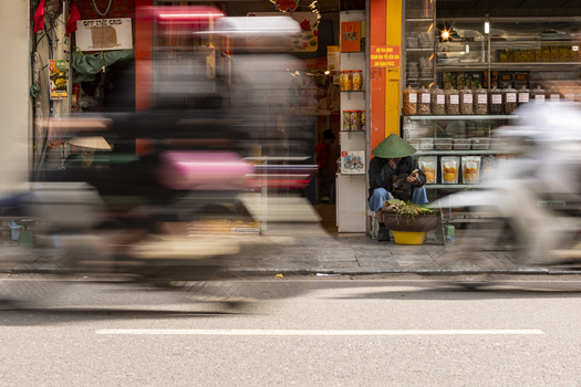 Hanoi Ha Noi Vietnam 11 November 2025 View of a person wearing a traditional conical hat sitting on the sidewalk in front of a shop while motorbikes pass by in a blur.