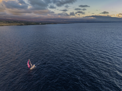Aerial view of a sailboat with a colorful sail navigating the deep blue waters of the Pacific Ocean near the coastline under a dramatic sunset sky in Waimea Hawaii United States. Aerial view of a sailboat with a colorful sail navigating the deep blue waters of the Pacific Ocean near the coastline under a dramatic sunset sky in Waimea Hawaii United States.