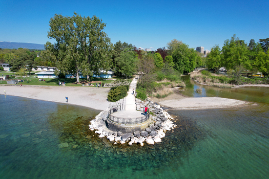 Aerial view of the stone pier and pebble beach surrounded by lush green trees and clear turquoise water of Lake Geneva in Morges Vaud Switzerland. Aerial view of the stone pier and pebble beach surrounded by lush green trees and clear turquoise water of Lake Geneva in Morges Vaud Switzerland.