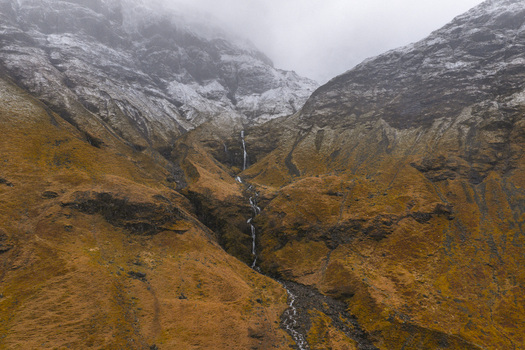 Aerial view of Glencoe valley with a thin waterfall cascading down rugged brown slopes under snow dusted peaks and misty clouds Glencoe Scotland United Kingdom. Aerial view of Glencoe valley with a thin waterfall cascading down rugged brown slopes under snow dusted peaks and misty clouds Glencoe Scotland United Kingdom.