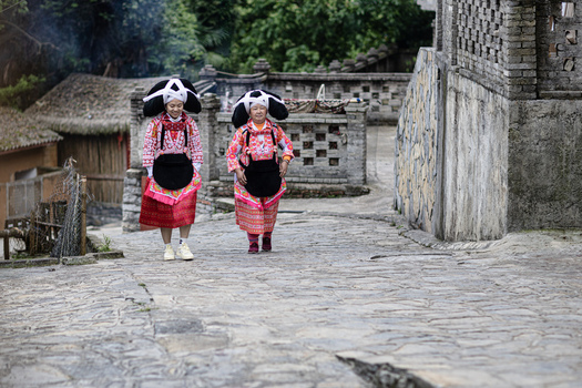 Gaoxing China 30 April 2026 View of two Long Horn Miao women in traditional ethnic costumes walking on a stone paved village street with stone walls and traditional houses. Gaoxing China 30 April 2026 View of two Long Horn Miao women in traditional ethnic costumes walking on a stone paved village street with stone walls and traditional houses.