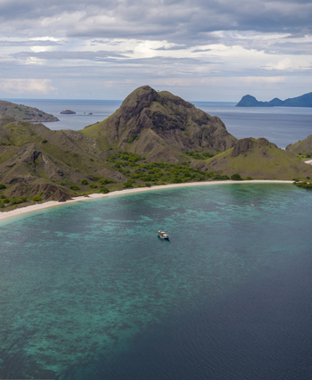 Aerial view of Komodo National Park featuring a small boat anchored in turquoise waters near a sandy beach and rugged hills in Komodo East Nusa Tenggara Indonesia. Aerial view of Komodo National Park featuring a small boat anchored in turquoise waters near a sandy beach and rugged hills in Komodo East Nusa Tenggara Indonesia.