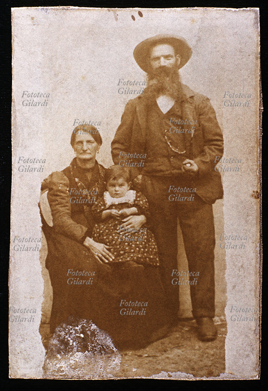 FAMIGLIA nonni in posa con la nipotina, il nonno in piedi fuma un lunga pipa e la nonna è seduta con in braccio la piccola. L\