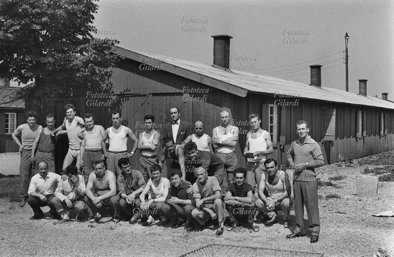 EMIGRAZIONE Italia Germania. Gruppo di lavoratori italiani fotografati davanti alle baracche adibite a loro abitazioni per scapoli, o per uomini senza famiglia al seguito.. Fotografia di Ando Gilardi (1921-2012) #andogilardi, Dachau, 1954 - 1955.