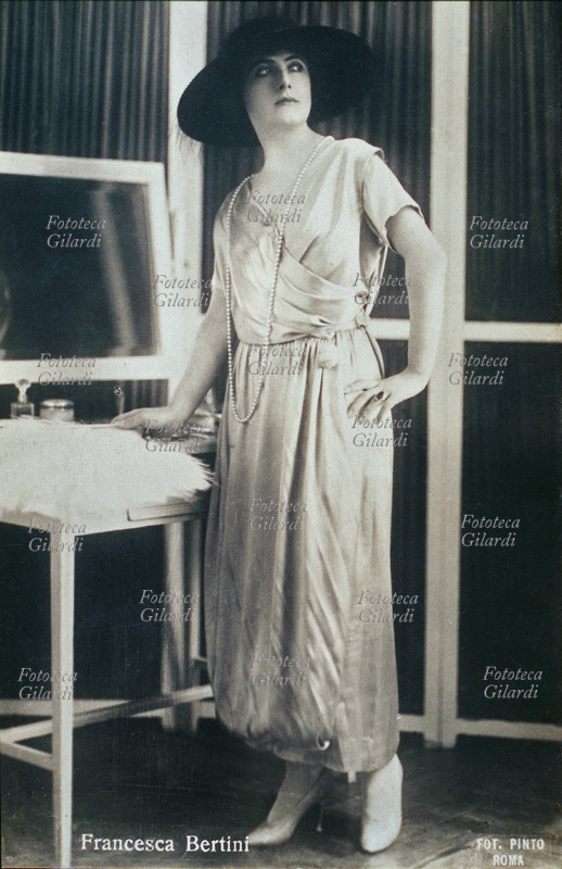 Francesca BERTINI (Elena Seracini Vitiello 1892-1985) attrice. Ritratto a figura intera, in piedi vicino ad una Toilette, con ampio cappello scuro. La sua fama di donna bella e fatale diede origine al divismo in Italia. Prese parte ad oltre 90 film, alcuni: Assunta Spina (1915), Fedora (1916), La donna nuda (1918). Fotografia Pinto, Roma 1915 circa.