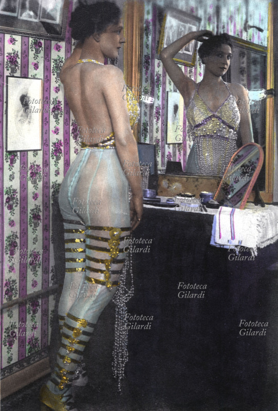 PROSTITUZIONE Fotografia di interno in una stanza del piacere. Lei si guarda allo specchio, indossa una calzamaglia trasparente decorata con nastri dorati. Primi anni del XX secolo