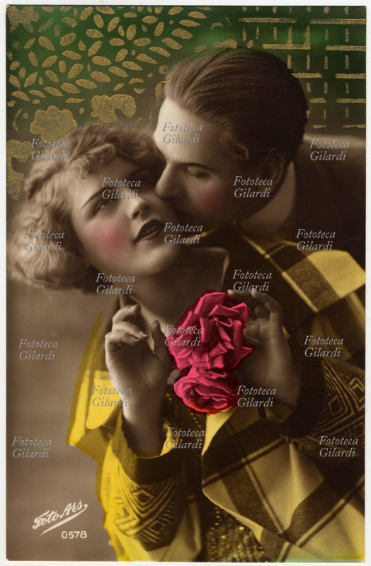 COPPIA Lui la bacia ardentemente sulla guancia, lei con lo sguardo sognante tiene in mano due rose rosa. Cartolina fotografica acquarellata e decorata sullo sfondo verde in oro. 1900 - 1930
