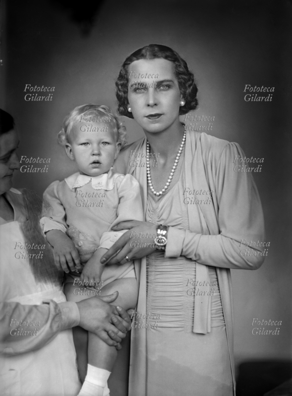 MARIA JOSÉ del Belgio (4 agosto 1906 – 27 gennaio 2001) Marie José Carlotta Sophia Amelia Enrichetta Gabriella di Sassonia Coburgo-Gotha, Principessa consorte del Piemonte in quanto moglie del futuro Umberto II dal 1930, con il secondogenito Vittorio Emanuele (12 febbraio 1937 – 3 febbraio 2024). La principessa è aiutata dalla balia nel sorreggere il piccolo, ritocco e taglio dell\