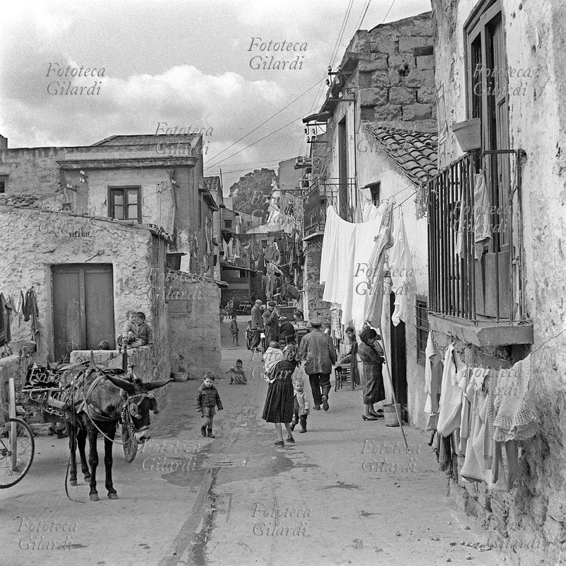 PALERMO vicolo affollato di adulti intenti al lavoro e bambini che giocano. Tanti sono i panni stesi che attraversano le case, un asinello stanco dorme attaccato al suo carretto. Fotografia di Ando Gilardi (1921 - 2012) #andogilardi da ampio servizio, Palermo, Cortile Cascino, 1957.