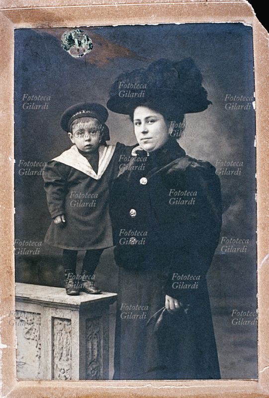 FAMIGLIA madre e figlio, entrambi elegantissimi, in posa dal fotografo. La donna indossa un vestito scuro con giubbetto di pelliccia e ampio cappello con piume. i guanti in mano; il bambino è vestito alla marinara e sul suo cappellino si intravede il nome \