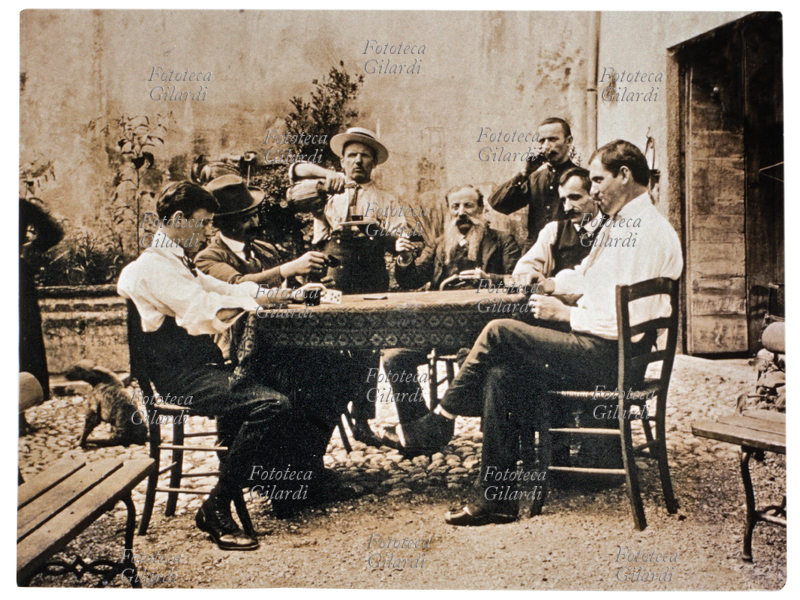 VINO Gruppo maschile ritratto a tavolino mentre gioca a carte. Sul tavolo insieme alle carte, sono disposti i bicchieri colmi di vino. Il nettare degli dei è indispensabile per la concentrazione nel gioco! Fotografia, Italia 1900 circa.