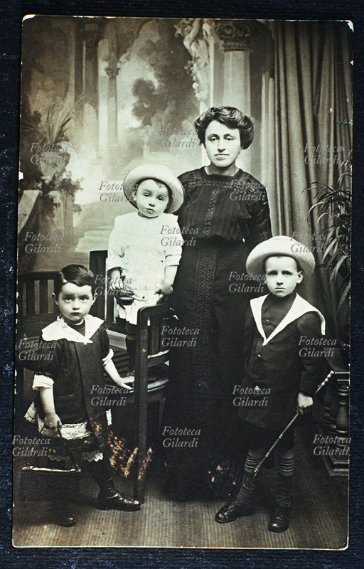 FAMIGLIA madre in posa con i tre figli, due maschi e una femmina. Sia i bambini che la donna indossano abiti che denotano la loro appartenenza ad una classe sociale benestante, i due più grandi vestono alla marinara, la femmina porta una borsetta e il maschio un bastoncino da passeggio; i due maschi indossano il cappello. Fotografia di anonimo eseguita in atelier, Italia 1900 circa.