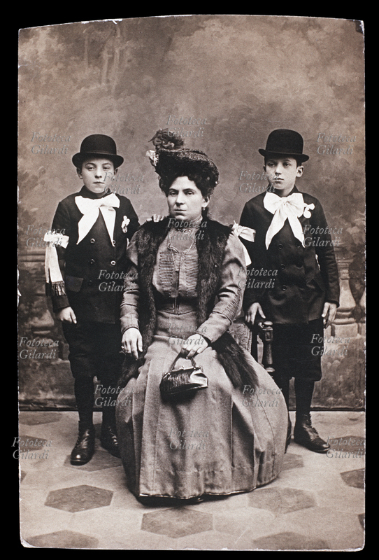 FAMIGLIA Fotografia ricordo della Cresima: due fratelli posano insieme alla madre. Lei, seduta al centro, indossa un vestito lungo con stola di pelliccia, cappellino alla moda e borsetta. I due ragazzi indossano il vestito della cerimonia con fiocco bianco al collo e al braccio e bombetta. Italia, 1900 circa