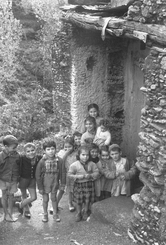 BAMBINI gruppo numeroso di diverse età, in posa accanto alla porta di un casolare in pietra sullo sfondo del bosco. Varie le espressioni: sorridenti, incuriosite e timorose. Fotografia di Ando Gilardi (1921 - 2012) #andogilardi, Nicastro 1955-58.