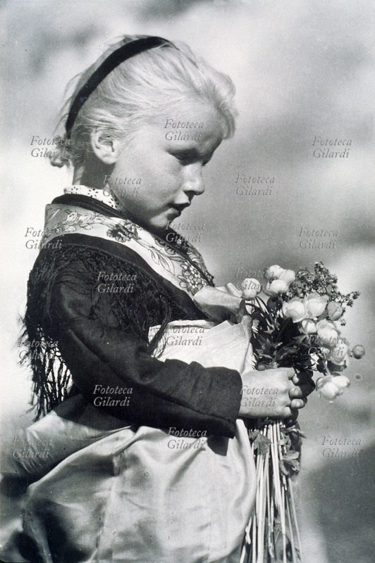 VALLE DI FUNES (BZ) bambina in costume tradizionale, ritratta di profilo, con un mazzo di fiori di campo. Fotografia, Wolfram Knoll, Bolzano 1959