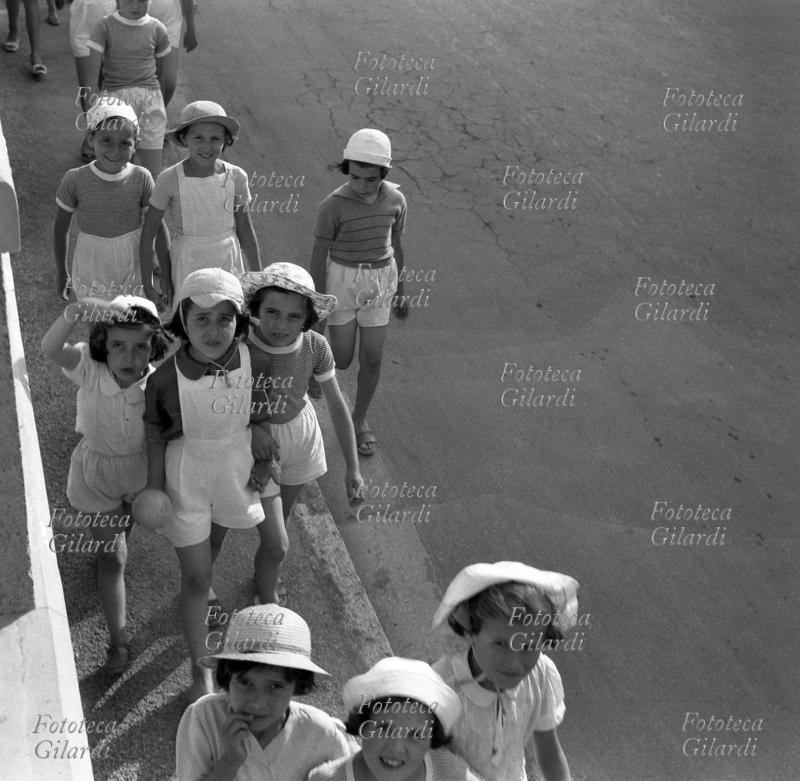 SCUOLA ESTIVA bambine in età di scuola elementare si avviano in fila per tre alla scuola estiva (detta anche colonia); indossano tuta o pantaloncini bianchi, una di loro ha in mano una palla. Fotografia di Ando Gilardi (1921 - 2012), #andogilardi, Roma, circa 1952.
