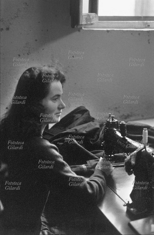 MESTIERI sarta alla macchina da cucire; la giovane sorride serenamente, ripresa di profilo al banco di lavoro posto sotto la luce della finestra, cuce con una delle prime \