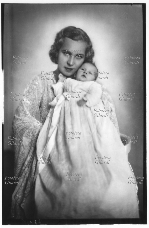MARIA JOSÉ del Belgio (4 agosto 1906 – 27 gennaio 2001) Marie José Carlotta Sophia Amelia Enrichetta Gabriella di Sassonia Coburgo-Gotha, Principessa consorte del Piemonte in quanto moglie del futuro Umberto II dal 1930, stringe teneramente la primogenita principessa Maria Pia (24 settembre 1934), che volge lo sguardo dritto e tiene la boccuccia leggermente aperta. La madre ha una espressione intensa e gioiosa. Ritratto fotografico di Ghitta Carell (20 settembre 1899 – 18 gennaio 1972), fotografa ufficiale dei reali d\