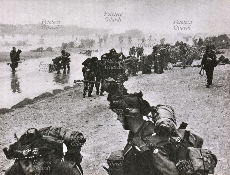 D-DAY Sbarco in Normandia - Sulle coste normanne sbarcano sempre più rinforzi: nella foto, soldati addetti al soccorso sanitario, riconoscibili dalla fascia sul braccio, si prendono cura di loro commilitoni, feriti in precedenza. Francia, 1944.
