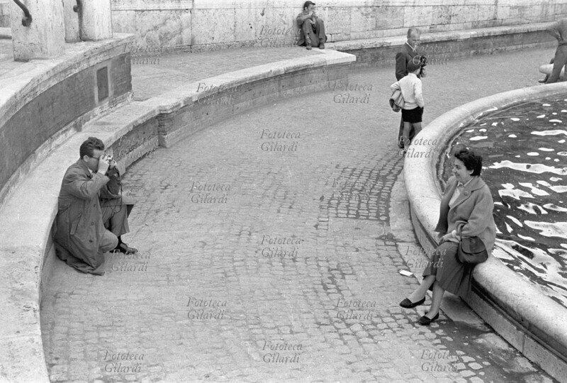 TURISMO coppia di turisti, lui fotografa lei seduta sul bordo della fontana di Trevi a Roma, in posa per una classica foto ricordo. Fotografia di Ando Gilardi (1921 - 2012) #andogilardi, Roma 1956