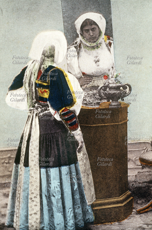 BONO (Sardegna) giovane donna in costume tradizionale, posa davanti ad uno specchio, per meglio evidenziare il suo abito particolarmente elaborato. Fotocollografia acquarellata, 1910 circa.