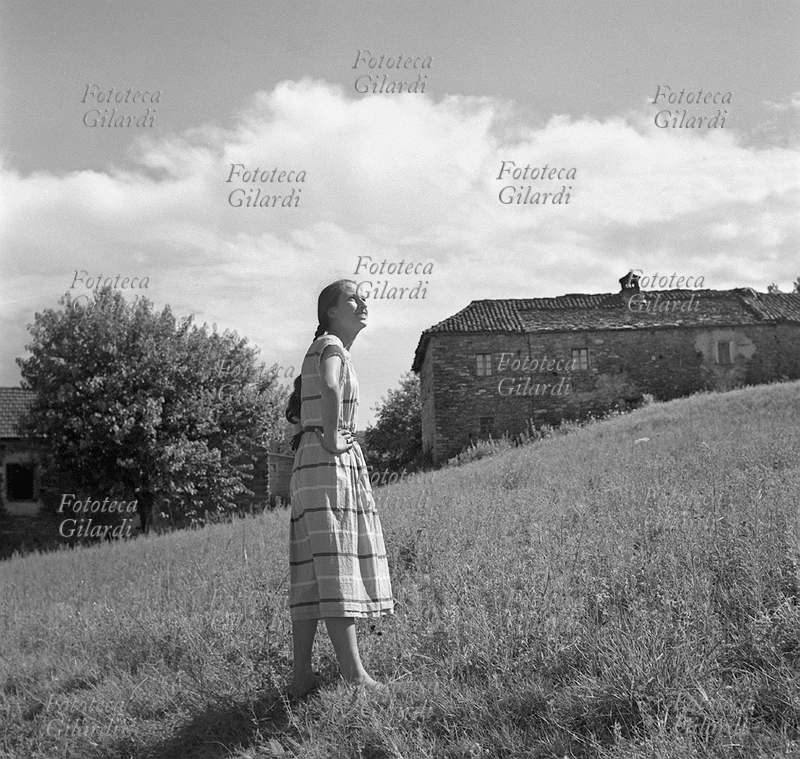 ADOLESCENTE ragazza ritratta in un campo, sullo sfondo di una cascina rurale tra le colline del basso Piemonte, in provincia di Alessandria. Fotografia di Ando Gilardi (1921 - 2012) #andogilardi, Italia 1952.