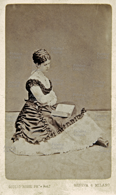 MODA Ritratto di giovane donna seduta in terra mentre legge un libro. Il ricco vestito lungo le copre le gambe, dalla gonna spunta appena uno stivaletto. L\