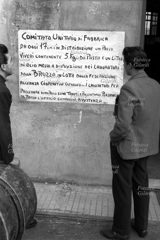 S.M.O. ITALIA lotte operaie alle Ferriere Bruzzo di Genova, in serrata da 35 giorni: operai leggono il cartello del Comitato Unitario di Fabbrica: la Federazione Alleanza Cooperative Genovesi, mise a disposizione di ciascuna famiglia un pacco di viveri contenente 5 Kg di pasta ed un litro d\