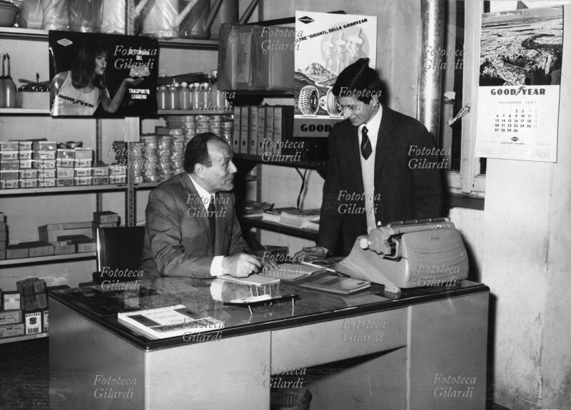 GOMMISTA ufficio del negozio-officina diagnosi, controllo e registrazione geometria avantreno vetture, in un quartiere periferico milanese. Il proprietario è in posa con il figlio. Milano, 1967.