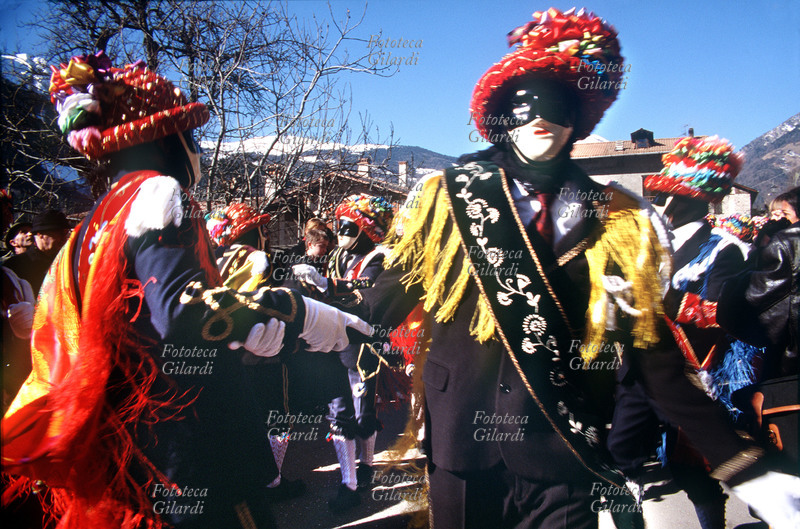 CARNEVALE Ballerini del Carnevale di Bagolino, durante una danza i ballerini indossano sontuosi indumenti rituali, composti da giacca e pantaloni al ginocchio di colore scuro, decorati con complessi ricami e passamaneria, spalline, alamari, gradi, tracolla con coccarda - tutti elementi di chiara derivazione militare - camicia bianca, cravatta, accessori, polsini in lana (manì), guanti in filo, calze bianche finemente lavorate, scarpe alte nere (polachì), ampio scialle in seta multicolore appuntato alle spalle, fascia ricamata sul braccio, foulard strettamente avvolto attorno al capo. I loro volti sono celati da maschere prive di espressione che assolvono un efficace ruolo di spersonalizzazione. Tuttora costruite da alcuni ballerini, esse sono realizzate in tela e gesso, internamente rivestite di cera e colorate di bianco, con bautta nera e labbra (talora anche le guance) rosse. L\
