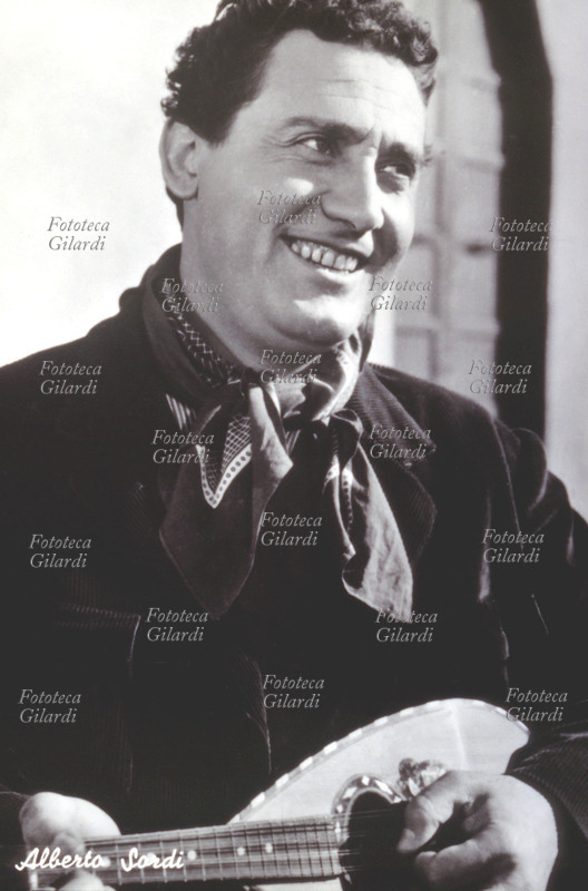 Alberto SORDI (15 giugno 1920 – 24 febbraio 2003), attore, regista, comico, sceneggiatore, conduttore radiofonico, compositore, cantante e doppiatore italiano. Ritratto mentre suona il mandolino in costume rurale per una fotografia promozionale diffusa in cartolina dalla casa cinematografica ad uso dei suoi fans. Italia 1952-59.