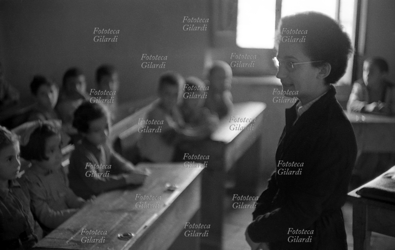 SCUOLA aula in classe mista, i bambini in età da scuola elementare (intorno ai sette anni) seguono la lezione dai banchi di fronte alla maestra. La classe è mista, ma divisa in due settori: a destra (guardando la cattedra) il femminile e a sinistra quello maschile. Fotografia di Ando Gilardi (1921 - 2012) #andogilardi, Melissa (Crotone) 1954.
