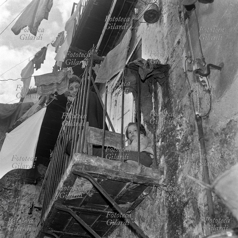 BAMBINI bambina sul balcone, insieme alla nonna, dietro alle sbarre della ringhiera che la rappresentano metaforicamente come in prigione. Il cielo nuvoloso aggiunge drammaticità alla situazione, sulla facciata della casa l\