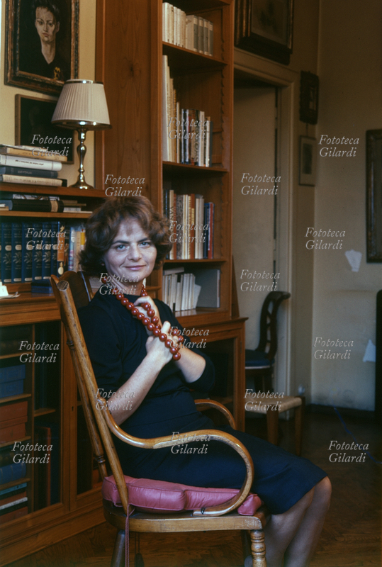 Elsa MORANTE (1912-1985) La scrittrice italiana, è ritratta seduta su una sedia in stile viennese, davanti ad un libreria. Indossa un abito blu scuro e una collana con grandi perle rosse a cui si aggrappa con le mani. ritratto fotografico realizzato da Ando Gilardi, s.d.