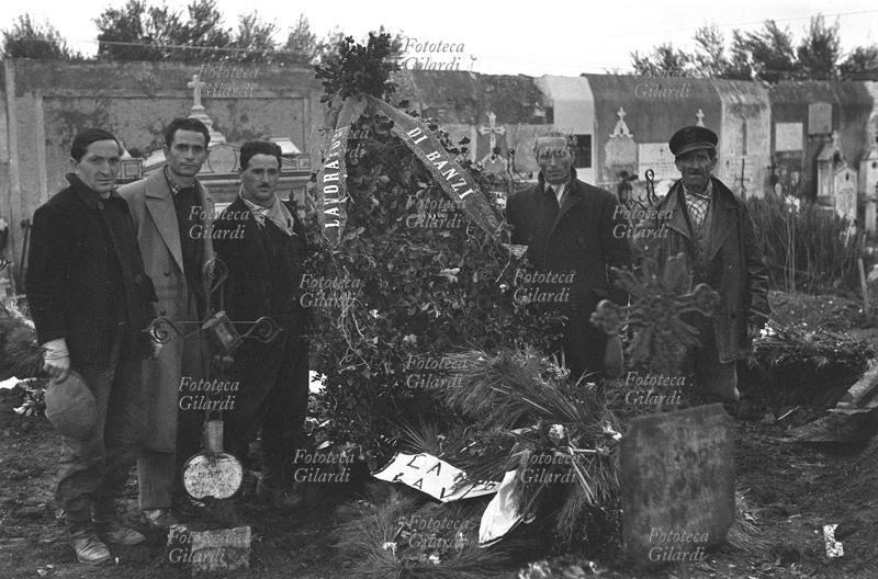 ITALIA funerali di Rocco Girasole ucciso dalla polizia il 13 gennaio 1956 durante lo sciopero a rovescio, una manifestazione di braccianti disoccupati che svolgevano volontariamente lavori pubblici di manutenzione della strada resa un fiume di fango dalle recenti nevicate; in questo modo evidenziavano rendendosi utili sia la propria inattività a causa della mancata promessa distribuzione delle terre, sia la lungaggine nei lavori di ripristino delle strade da parte delle autorità preposte. La tomba di Girasole; i camposantari e alcuni dirigenti locali del PCI in posa accanto alla corona dei lavoratori di Banzi. Fotografia di Ando Gilardi (1921 - 2012) #andogilardi, parte di un ampio servizio realizzato per \