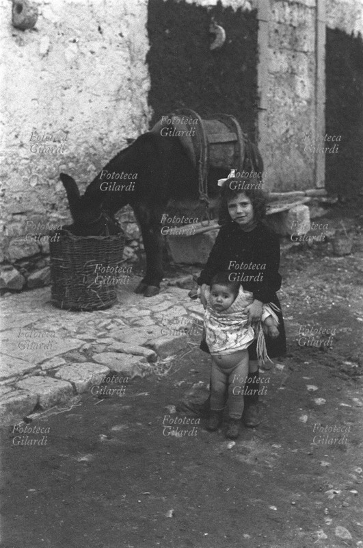 ITALIA Venosa, bambine davanti ad una abitazione, la più grandicella bada alla più piccina; sullo sfondo, il mulo imbastato mangia in una cesta. Fotografia di Ando Gilardi (1921-2012) #andogilardi, parte di un ampio servizio realizzato per \