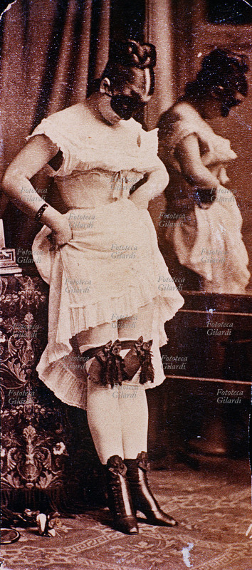 EROTISMO Giovanetta in biancheria intima solleva la sottogonna e mostra, guardandole, le giarrettiere ornate di due grosse gale. La fanciulla si nasconde il viso con una mascherina nera, e la sua immagine è riflessa in profilo nello specchio. Fotografia fine del XIX secolo.
