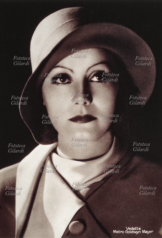 Greta GARBO (1905-1990) ritratta in primo piano frontalmente con un cappello di feltro e cappotto. Immagine fotografica pubblicitaria distribuita da Metro Goldwin Mayer 1930 circa