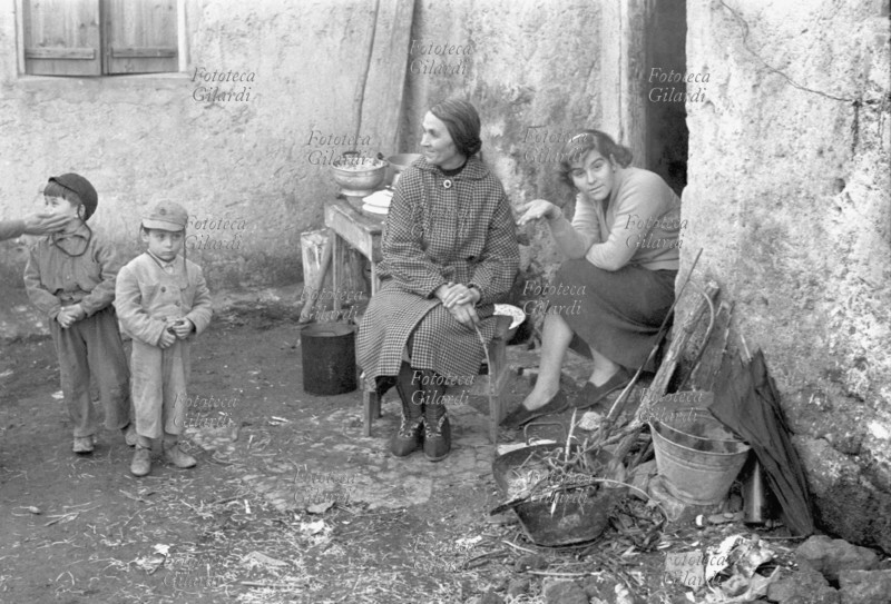 FAMIGLIA. Donne dei sobborghi romani ritratte sulla soglia della loro abitazione. Una donna, seduta su una sedia, indossa un soprabito a quadretti. Una ragazza, seduta sulla soglia, sorride all\