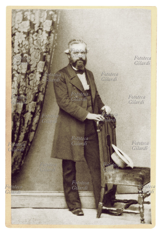 Karl MARX (1818-1883) in piedi appoggiato ad una seggiola su cui è posato un cappello. Ritratto fotografico di anonimo, fotografia realizzata in studio, Germania 1879 circa