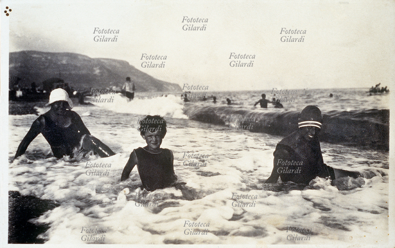 VACANZE MARE bambino e due ragazze in posa sul bagnasciuga tra i flutti del mare grosso. Il controluce, la notevole abbronzatura dei soggetti ed il contrasto con la schiuma bianca, producono l\