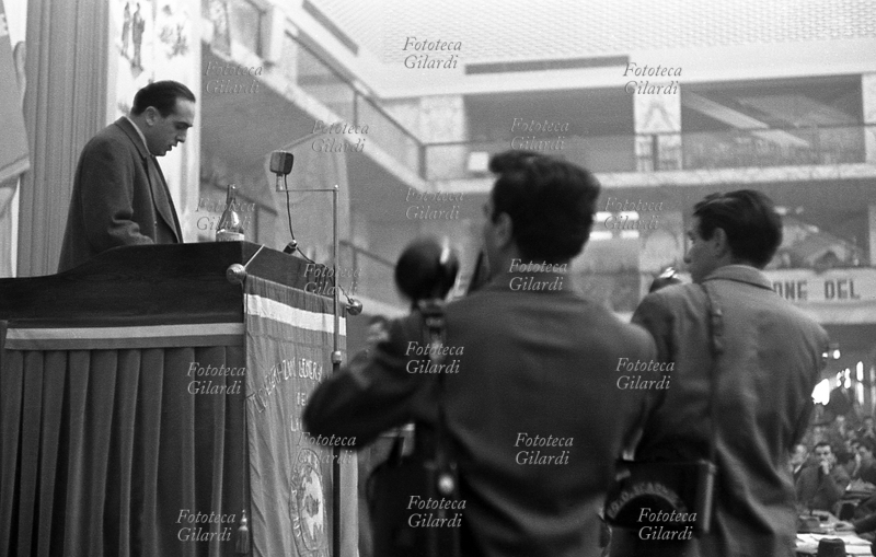 IV CONGRESS of the CGIL meeting in Rome EUR. A speaker on stage speaks to a packed room, filmed by two photographers; on the podium the microphone for amplification and the one for recording. Photograph by Ando Gilardi (1921 - 2012), #andogilardi, Rome, 27 February/4 March 1956.