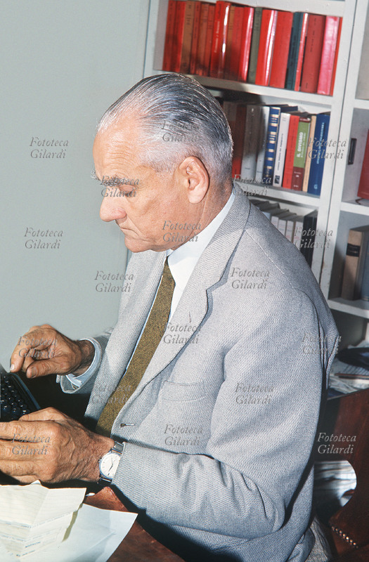 Alberto MORAVIA (Alberto Pincherle 1907-1990), scrittore, al lavoro nel suo studio, guarda in basso verso la macchina da scrivere. Ritratto fotografico eseguito da Ando Gilardi (1921-2012). Italia, 1957 circa