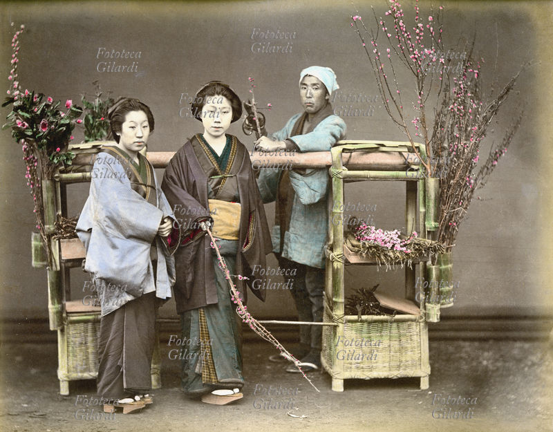 GIAPPONE 1890 Due donne ritratte insieme ad un venditore ambulante di rami fioriti di pesco, periodo Meiji (1868-1912). Fotografia colorata a mano, 1890 circa.