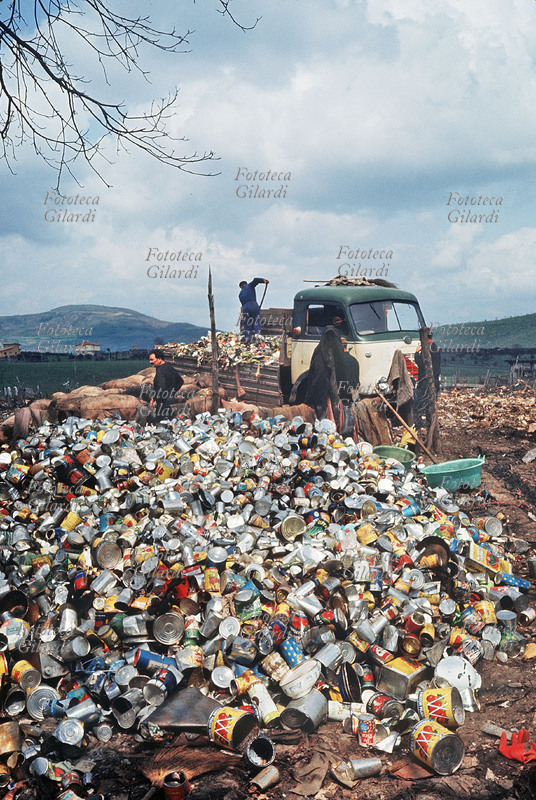 SPAZZATURA In una discarica alla periferia di Roma si utilizzano i rifiuti organici per allevare i maiali; i materiali non commestibili come lattine, stoffe, vetri, legname vengono recuperati e riciclati. Seppure di quantomeno dubbia liceità, si tratta di uno dei primi utilizzi economici della spazzatura proveniente dalla città, raccolta in maniera indifferenziata. Il porcaro bada ai maiali mentre altri due addetti si occupano del carico di un camioncino. Fotografia di Ando Gilardi (1921-2012) #andogilardi. Italia, 1955 circa.