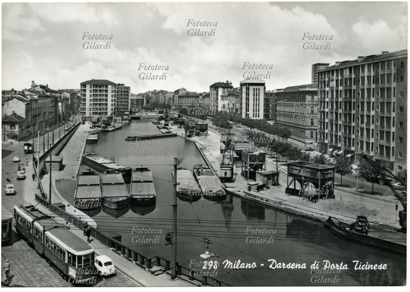 MILANO Darsena di porta ticinese con le chiatte e le gru per il carico/scarico delle merci nel passaggio da trasporto fluviale a trasporto su strada. In piena ricostruzione post-bellica, nel 1953 la darsena di Porta Ticinese è al tredicesimo posto nella classifica dei porti nazionali per ricevimento merci. Cartolina fotografica, Milano 1953